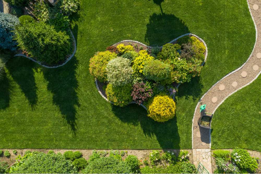 An aerial view showcases a landscaped garden with a heart-shaped flower bed of various green and yellow plants, embraced by a winding stone path. A person in a green top pushes a wheelbarrow along the path, perhaps attending to tree maintenance tasks throughout the serene scene.