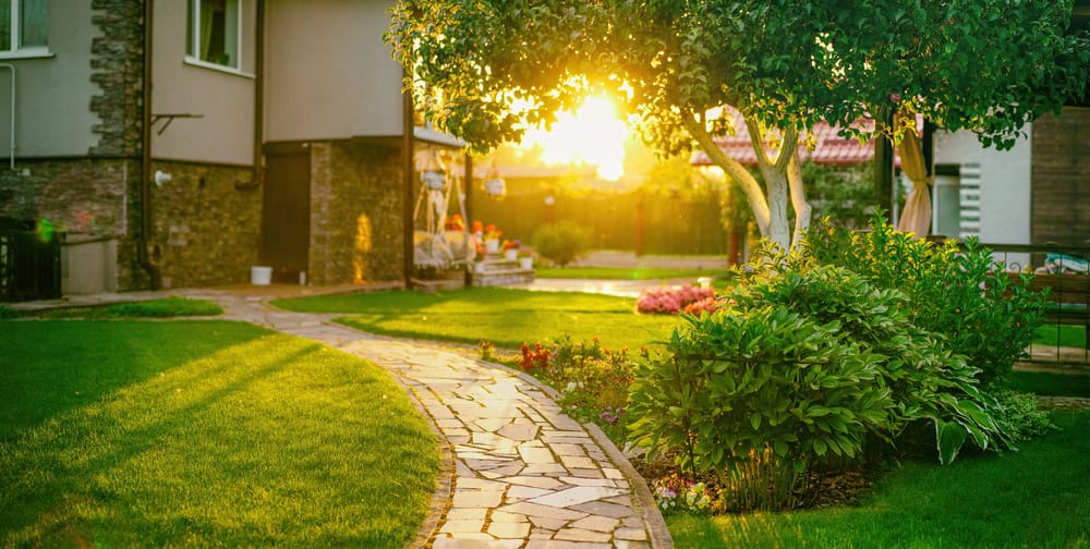 A serene garden path winds through lush green grass, flanked by vibrant plants and trees. The golden glow of a setting sun casts long shadows, creating a warm and inviting atmosphere in the tranquil backyard setting.