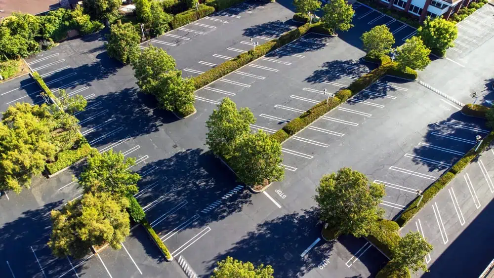 Aerial view of an empty parking lot with green trees interspersed between parking spaces. The lot is divided by landscaped sections and surrounded by greenery, with clear parking lines on a smoothly paved surface.