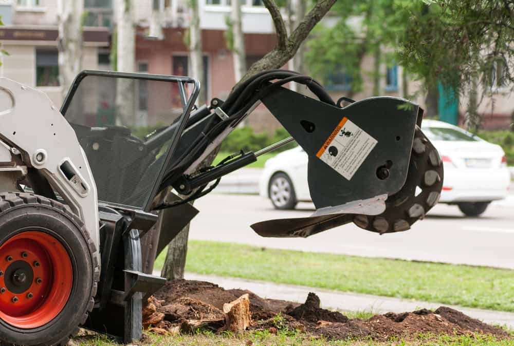 A tree stump grinder machine with a large rotating blade is positioned over a tree stump on a grassy area by the roadside. The background shows a blurred street with parked and moving cars.
