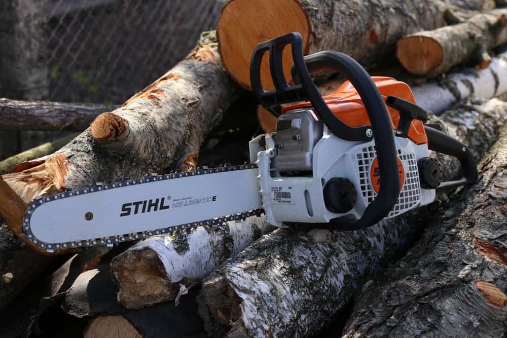 A Stihl chainsaw rests on a stack of cut logs. The chainsaw features an orange and white body with a metal blade, set against a background of outdoor logs with bark.